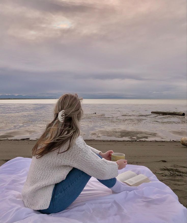 a woman sitting on top of a white blanket next to the ocean holding a drink