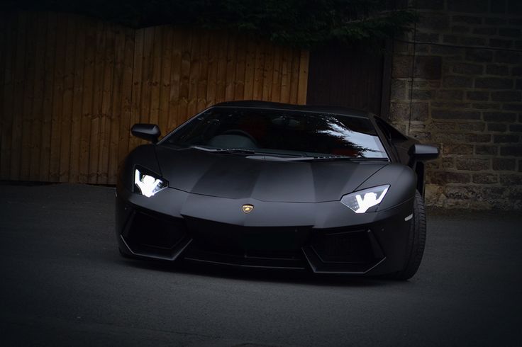 a black sports car parked in front of a wooden fence with its headlights turned on