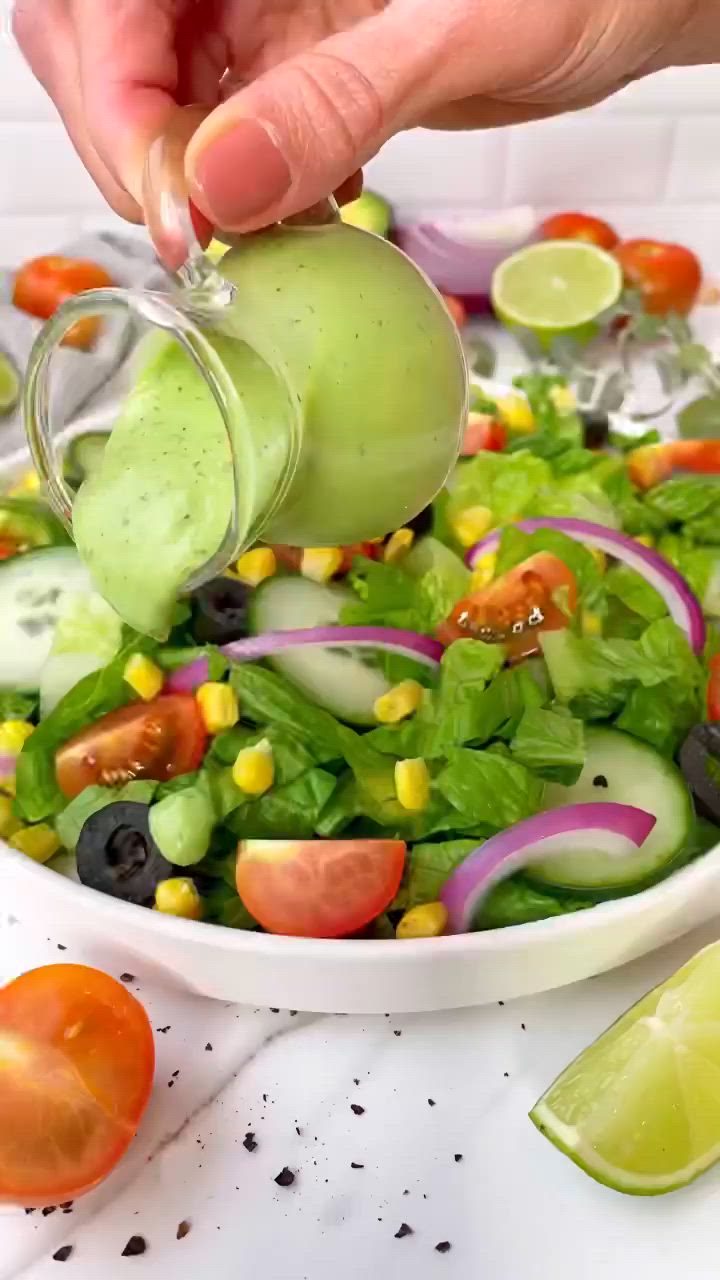 This may contain: a person pouring dressing into a salad in a white bowl with tomatoes, lettuce, corn and black olives