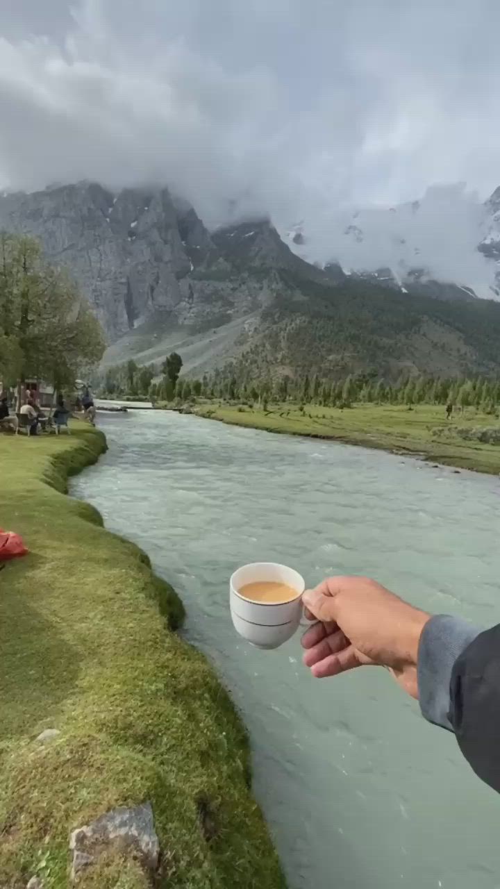 This may contain: a person holding a cup of coffee in front of a body of water with mountains in the background