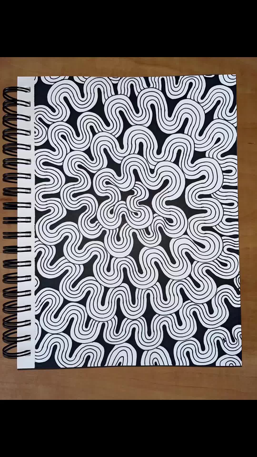 This may contain: a spiral notebook on a wooden table with black and white paper in the middle that has an abstract design