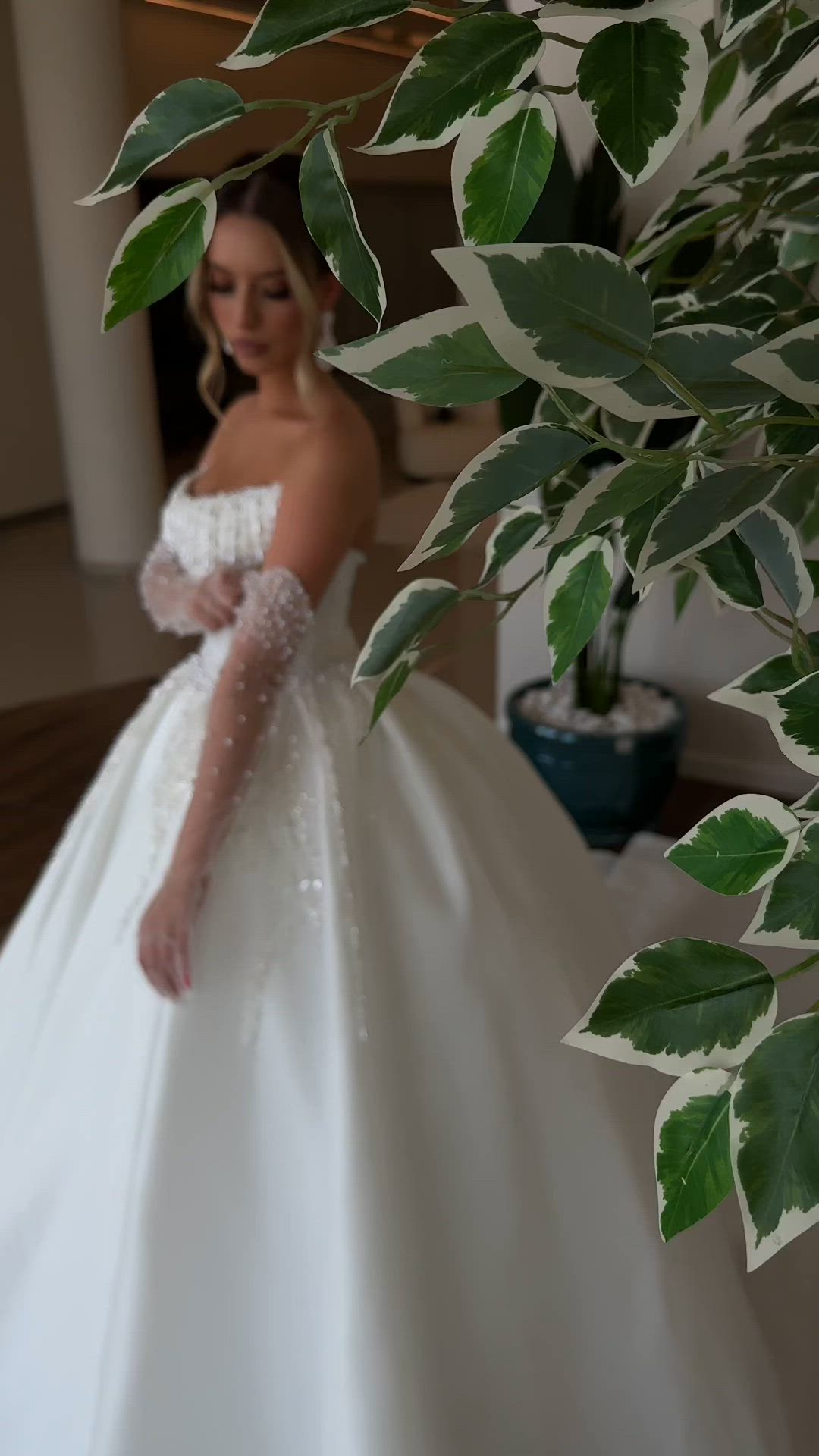 This may contain: a woman in a white wedding dress standing next to a tree with green leaves on it