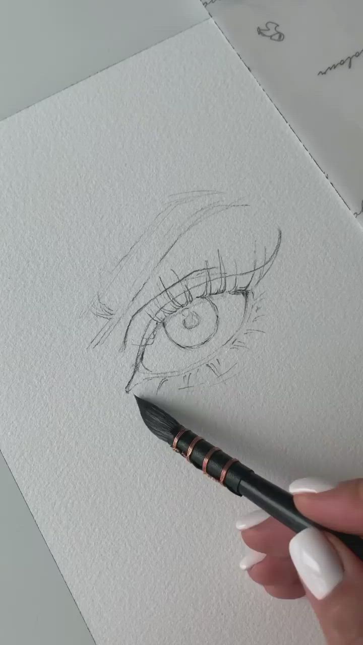 This may contain: a woman's hand is holding a pencil and drawing an eye on white paper