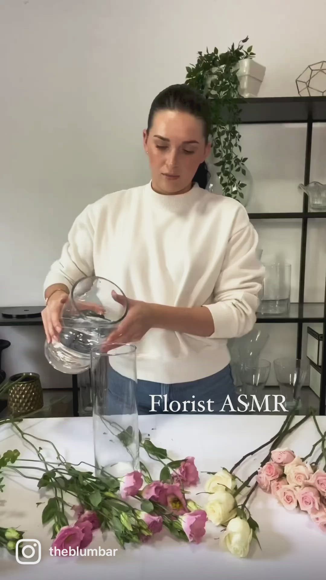 This may contain: a woman pouring water into a blender with flowers on the table in front of her