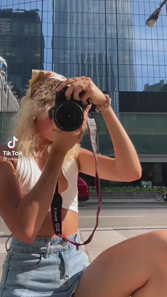 This may contain: a woman sitting on the ground with a camera in her hand and buildings behind her