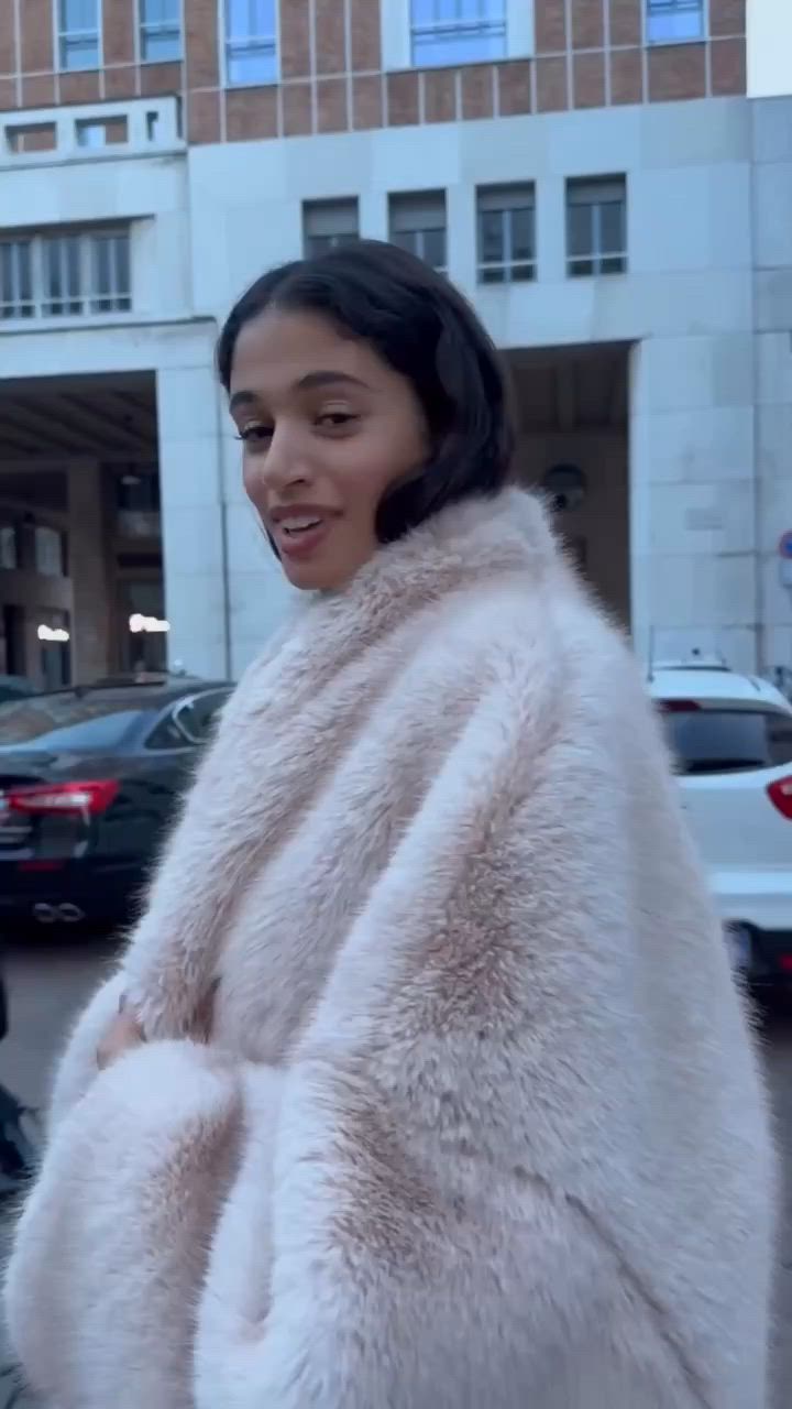 This may contain: a woman in a white fur coat standing on the street smiling at the camera with her hands under her chest