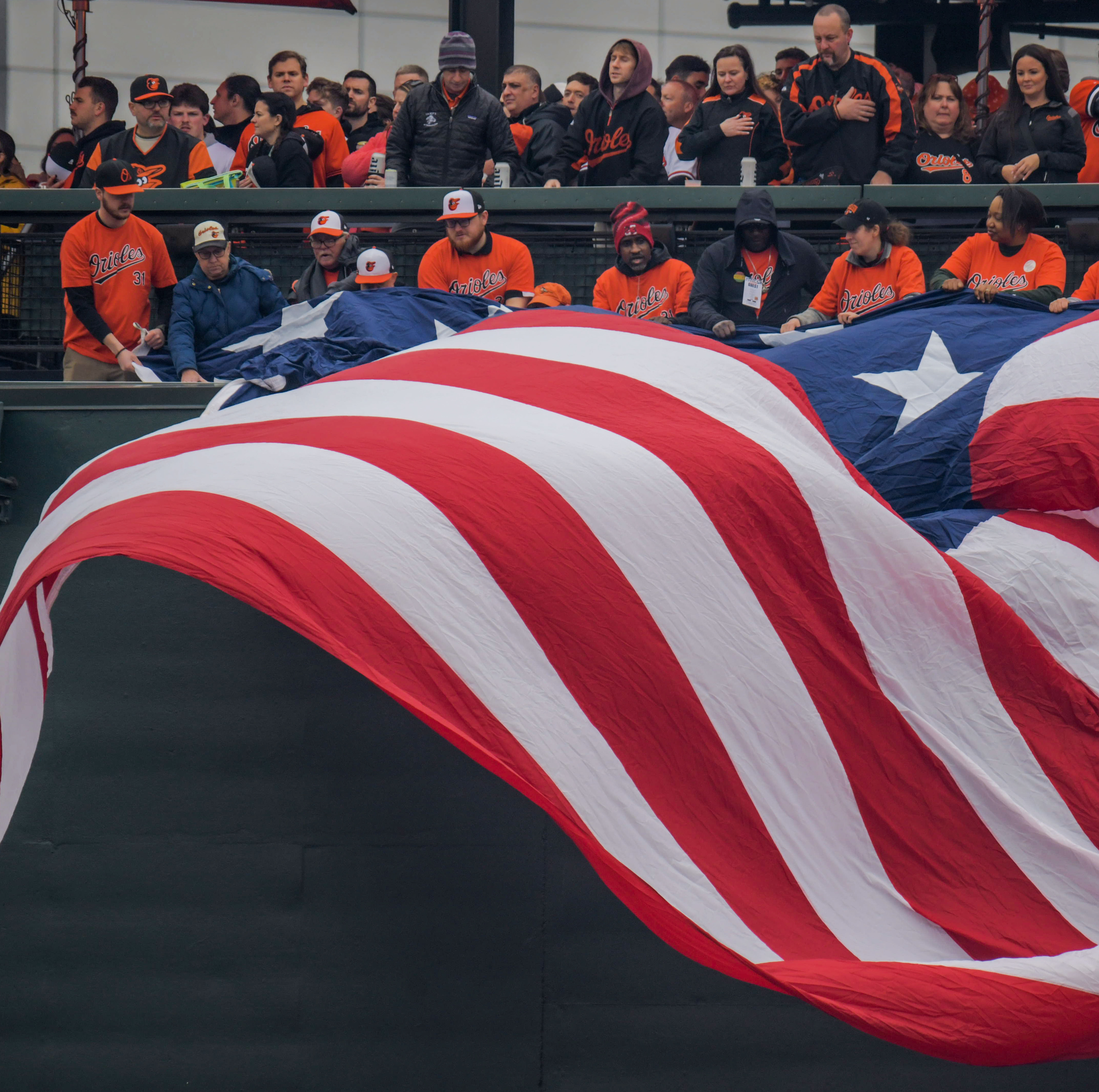 Mar 28, 2024: Attendees help unfurl a replica flag of Fort McHenry honoring the Francis Scott Key Memorial Bridge disaster during opening day of Major League Baseball at Oriole Park at Camden Yards. (Karl Merton Ferron/Staff)
