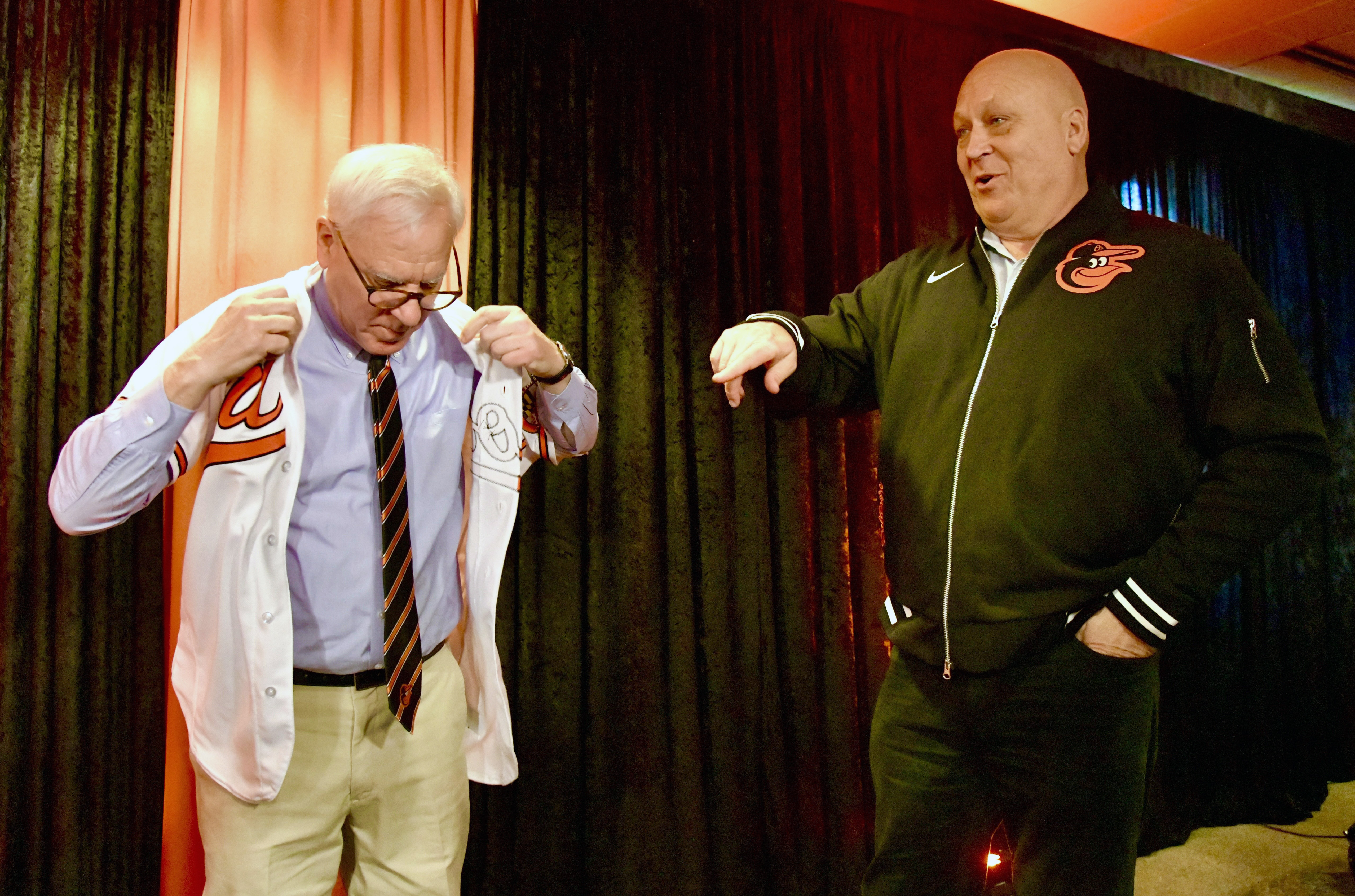 March 28, 2024: David Rubenstein, new owner of the Baltimore Orioles, tries on a team shirt with his name on the back, as Cal Ripken Jr., part of the ownership group, looks on during a news conference on opening day. (Amy Davis/Staff)