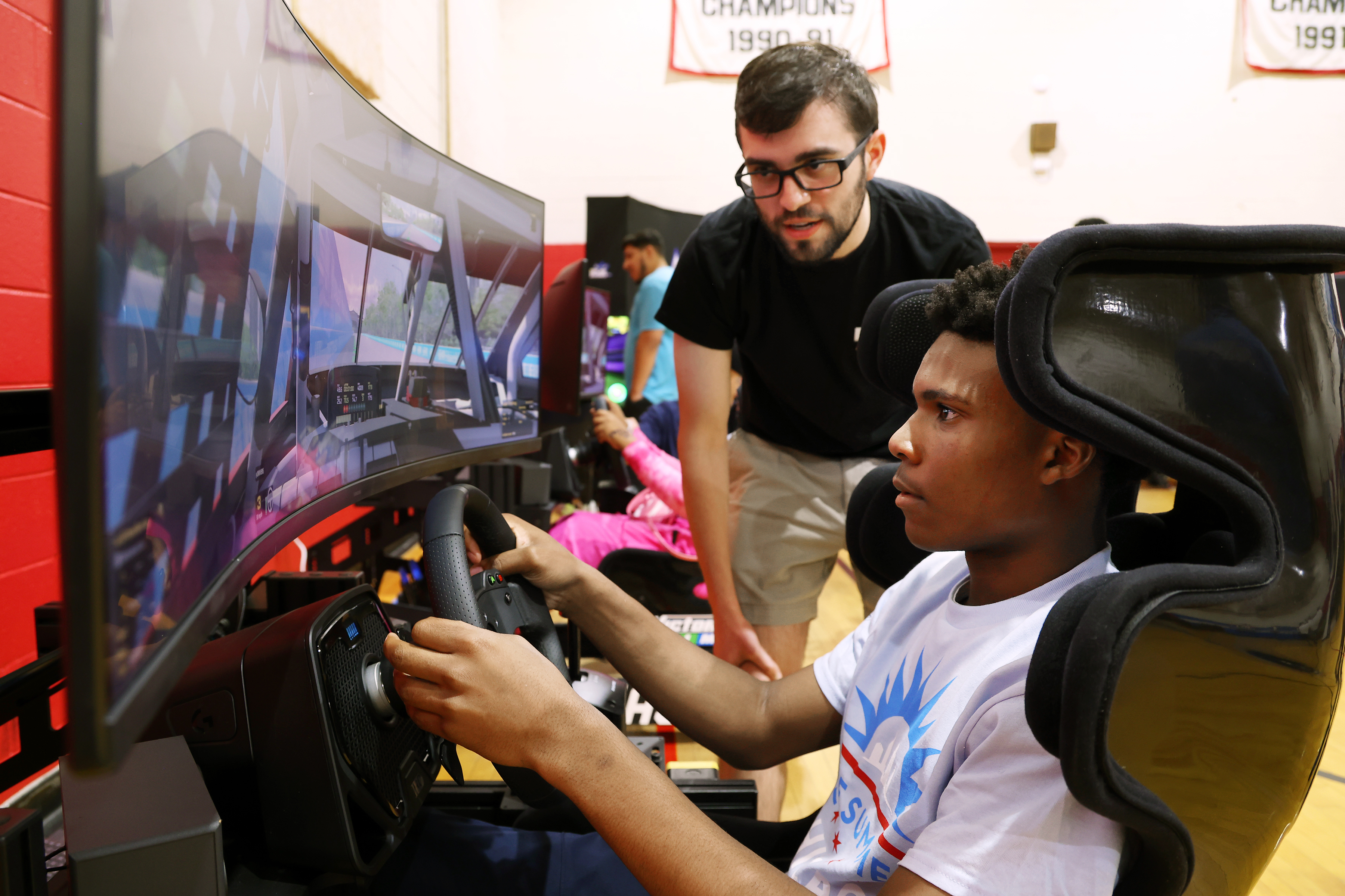 Jordan Coleman, age 14, tries out the iRacing gaming experience, an authentic auto racing simulator, at a NASCAR-themed event in the James Jordan Boys & Girls Club in Chicago on Tuesday, July 2, 2024. Ty Burress, rear, who set up the simulator, gives Coleman driving tips. (Terrence Antonio James/Chicago Tribune)