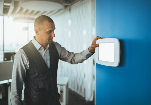 A mature handsome caucasian man entrepreneur using a multifunctional wall-mounted terminal panel with a blank white screen mock-up to adjust illumination level in the office and indoor air temperature