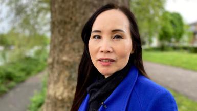 A woman in a blue coat standing in front of a tree in a park looking at the camera.