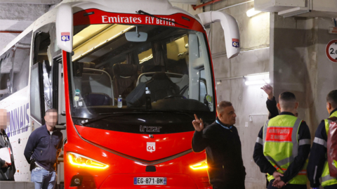 Firefighters stand next to Lyon's team bus, showing one window (left) completely broken and another damaged