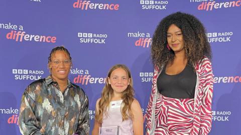 Harli, a 10 year old girl, holds an award trophy. She is standing between the awards host Angelle Joseph (left) and first round judge Lily Hammond (right)