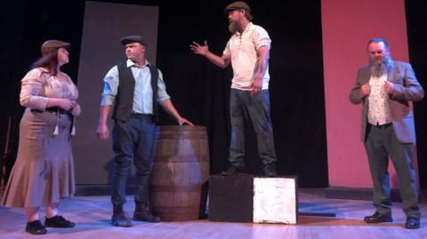 Four actors on stage performing a scene from the play Iniquity, which tells the story of Dic Penderyn. An actor in a white shirt and flat cap is standing on a box gesturing towards a male and female actors to his right. Another actor is standing on his left clutching his blazer.