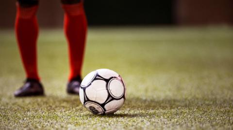 A green football pitch. In the centre there is a football. Behind the football are two legs, from the knees down, wearing red knee high socks and black football shoes. 