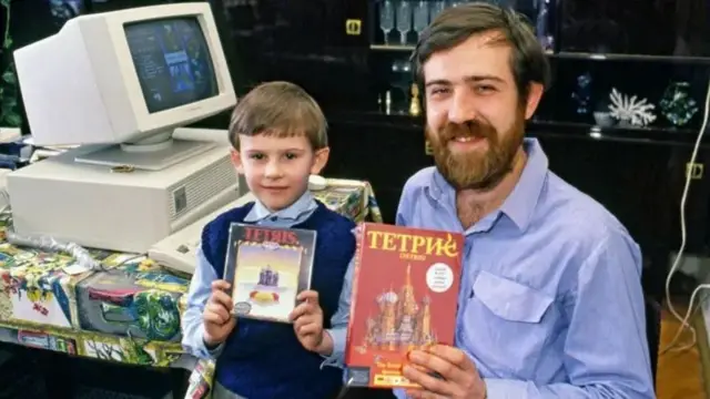 Alexey e o filho pequeno sorrindo e mostrando caixa de Tetris, perto de computadorAlexey Pajitnov and a boy holding Tetris boxes. in front of a 1980s computer