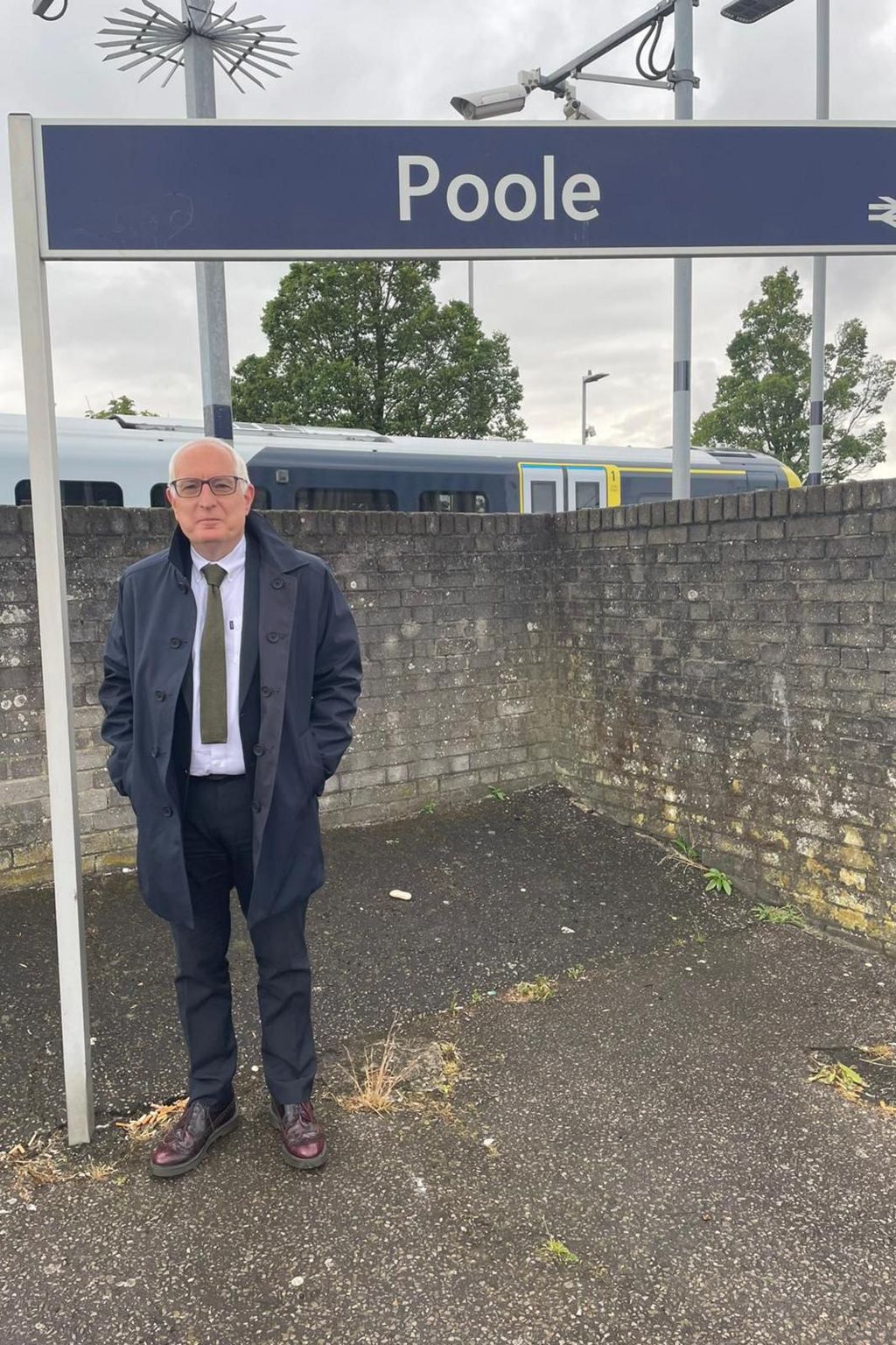 Neil Duncan-Jordan standing in front of the Poole sign at the railway station. he is wearing a dark suit, shite shirt and grey tie. He also has on a coat and brown shoes. He is clean shaven, has short white hair and is bald on top. He is also wearing glasses