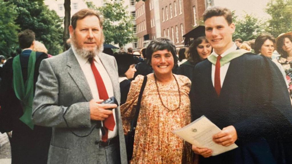 A smiling Keir Starmer wears a graduation gown and holds a certificate. His parents stand beside him.