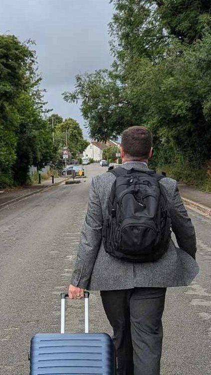 Back of Sean Woodcock walking down a road with a wheeled suitcase