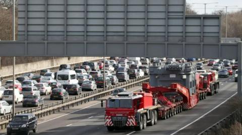 A long, wide, red lorry carrying a huge transformer travels down a very busy motorway 