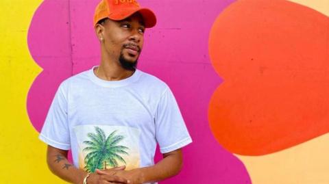 Reggie Lambe looks away from the camera wearing a white t-shirt and orange baseball cap