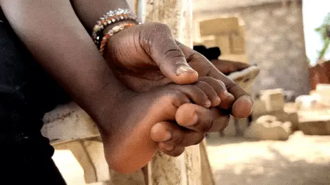 Image of a baby's foot, which is being held in the hand of its mother. There are concrete buildings in the background and the sun is shining