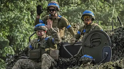 Three Ukrainian soldiers sit on top of tank in Sumy region wearing helmets and military gear with blue stripes, on 11 August