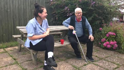 Chestnut Lodge resident Ted and carer Madi sit at a bench enjoying a cup of tea in the garden. 