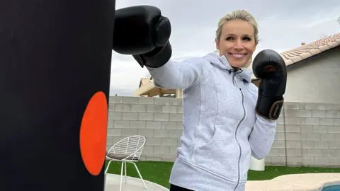 Lara Lewington wears black boxing gloves and punches a punchbag with a orange spot on it.