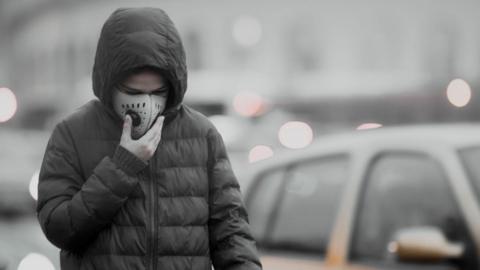 Boy in grey jacket, walking by vehicles clutches his face mask