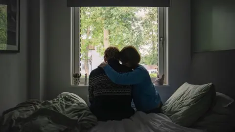 Getty Images A posed picture of two adults, a man and a woman, sitting on a bed at a window. The woman is hugging the man as he looks out the window