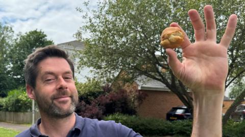 Man holding a scone-like round biscuit in his hand. Looking at the camera. In a small green field. He is wearing a navy top.