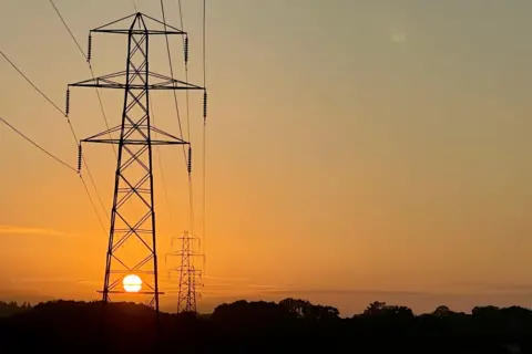 The rising sun shines orange through the metalwork of an electricity pylon which rise above the silhouette of trees on a low horizon. The wide expanse of sky is illuminated with a graduating orange emanating from the early morning sun.