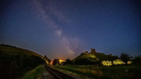 FRIDAY - The night sky over the Dorset countryside. A railway line runs through the centre, with the ruins of Corfe Castle visible on a hill to the right with a green hill to the right of the tracks It is night so the colours are muted, but in the distance you can see the glow from the street lights in Corfe Castle village. In the dark sky above there are thousands of stars with the milky way running through the centre of the sky.