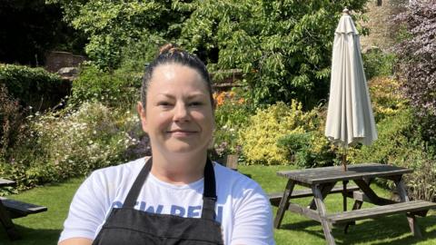 Pub owner and chef wearing black apron standing in beer garden surrounded by flowers