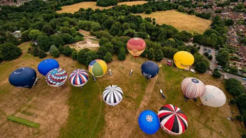 An aerial image of 13 colourful hot-air balloons about to take off from a field