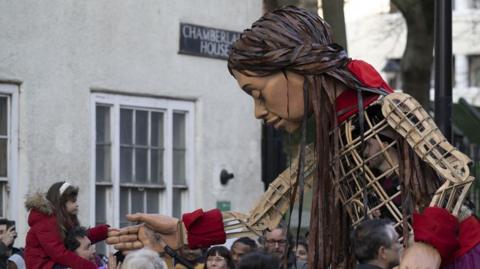 Little Amal, a large puppet with dark skin and hair, shakes hands with a young girl perched on a man's shoulders 