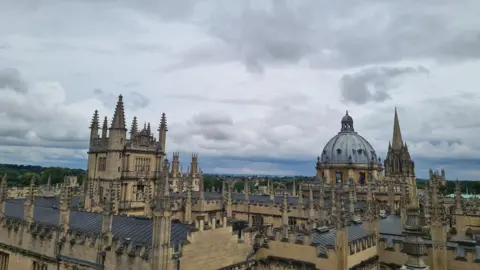 Rebekah WEDNESDAY - The roofs of Oxford are photographed from roof height against a grey sky. The buildings are made from a yellow stone with grey roofs. Each building has several stone spires. In the distance, the domed roof of the Radcliffe Camera looks striking. On the horizon you can see green trees and overhead the sky is full of light grey clouds.