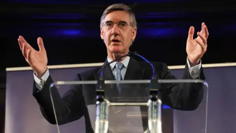 Jacob Rees Mogg standing in front of a glass podium, opening his arms and hands as he addresses an audience through a microphone. He is wearing a black jacket, white shirt and a silvery-coloured tie