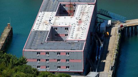 Bibby Stockholm, a grey, rectangular barge moored against a dock. It shows 12 windows.