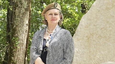 Joanna Burri-Weaver wears a military beret and stands next to a stone memorial in a forest