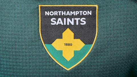 New Northampton Saints crest which is black and green with a gold cross across the middle