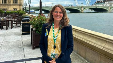 Vikki Slade stands outside with the London Eye in the background and a bridge over the River Thames. She is smiling at the camera, has mid length light brown hair and wears a navy blue blazer with a yellow and white floral print top underneath. 