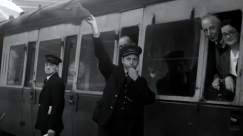 Black and white archive footage of a train full of people leaving a platform. A man is stood on the platform blowing a whistle and waving some fabric. Passengers can be seen looking out of the windows. 