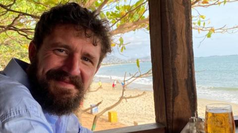 A smiling Simon Boas sitting in a beach hut with a drink in front of him on a table