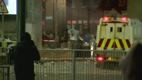 a man with his face covered running alongside a PSNI vehicle; he looks like he is about to throw a projectile that he is holding