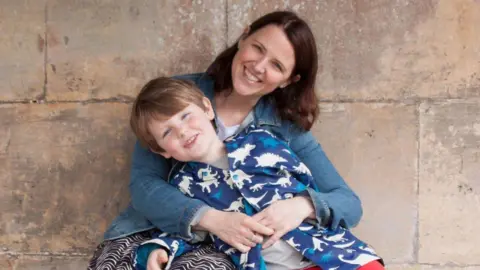 Rachel Gibson with shoulder length dark hair wearing a denim shirt with her arms around her son, aged about eight or nine, wearing a blue top with white and bright blue dinosaurs, sitting against a stone wall