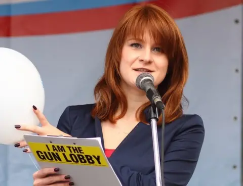 Civic Chamber of the Russian Federation Butina speaks into a microphone while holding a clipboard which says, on its back: I am the gun lobby