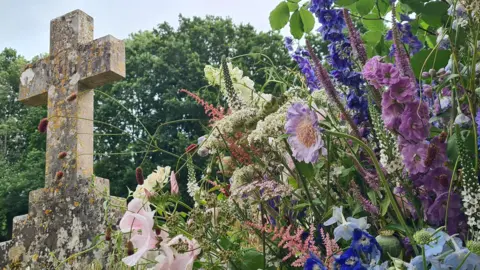 Rebekah THURSDAY - Wildflowers growing in a graveyard in Appleton. On the right of the frame are pink, purple and white flowers with green leaves. To the left is a large stone cross with yellow moss growing on it. To the rear are several large trees and overhead the sky is grey