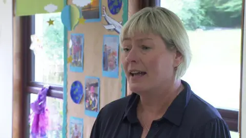 BBC Carol Shaw has short blond hair and is wearing a purple shirt. She's standing in a classroom with pupil's artwork pinned on the wall.